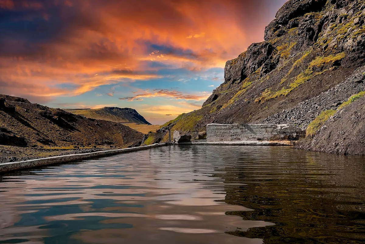 Source chaude de Seljavallalaug : Sérénité au coucher du soleil Un coucher de soleil paisible baigne les montagnes escarpées et les eaux tranquilles de la source chaude de Seljavallalaug dans des couleurs chaudes. Le reflet sur la surface calme ajoute à la beauté surréaliste du paysage islandais.