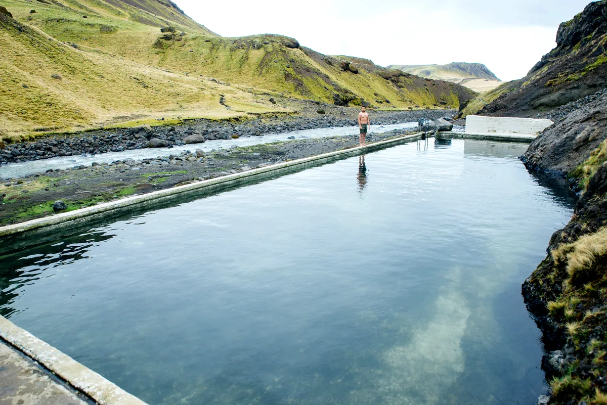 Piscine géothermique de Seljavallalaug : l'oasis cachée de la nature Un homme se tient près du bord de la source géothermique claire de Seljavallalaug piscine, entourée de collines couvertes de mousse. Le paysage naturel offre une évasion sereine et isolée en Islande.
