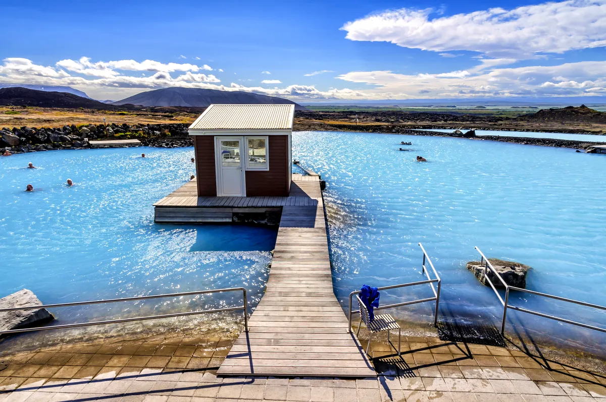 Myvatn Spa par temps clair Lagon géothermique de Myvatn sous un ciel clair, avec des eaux bleu vif reflétant la lumière du soleil. L'image capture le petit bâtiment central entouré de piscines, avec des montagnes et un vaste paysage pittoresque en arrière-plan.