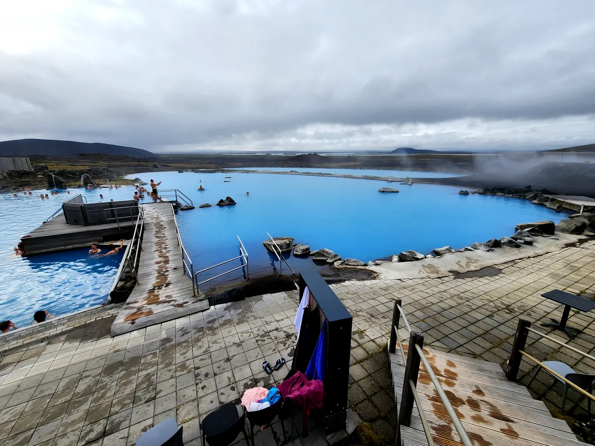 Le Blue Lagoon géothermique de Myvatn Une vue panoramique du spa géothermique de Myvatn en Islande, avec ses vastes piscines bleues se fondant dans l'horizon brumeux. Des plates-formes en bois entourent les piscines, offrant aux visiteurs une expérience immersive et relaxante. Le paysage environnant est accidenté, mais serein, offrant un contraste saisissant avec les eaux bleues apaisantes.