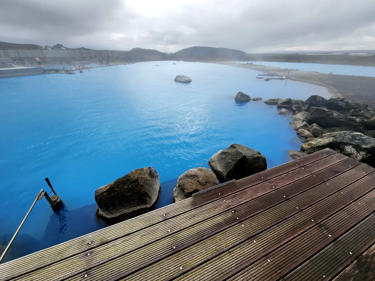 Les eaux sereines du lagon de Myvatn Une vue rapprochée des eaux bleues calmes du lagon géothermique de Myvatn en Islande. Les rochers et les passerelles en bois créent une atmosphère paisible tandis que la vapeur s'élève des eaux chaudes, tandis que le paysage lointain, enveloppé de brume, ajoute une touche mystérieuse au décor.