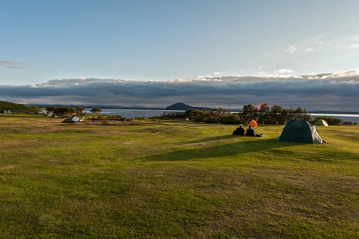 Un camping pittoresque en Islande avec des tentes installées sur une herbe verdoyante, surplombant un lac paisible et des montagnes au loin—illustrant la beauté des campings aménagés pour les voyageurs en van.