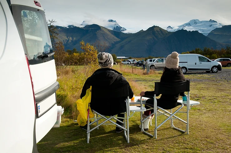 Deux voyageurs profitant d’un camping pittoresque en Islande, assis à l'extérieur de leur van avec une vue sur les montagnes et les glaciers. Un exemple parfait d’un site de camping bien équipé pour les voyageurs en van.
