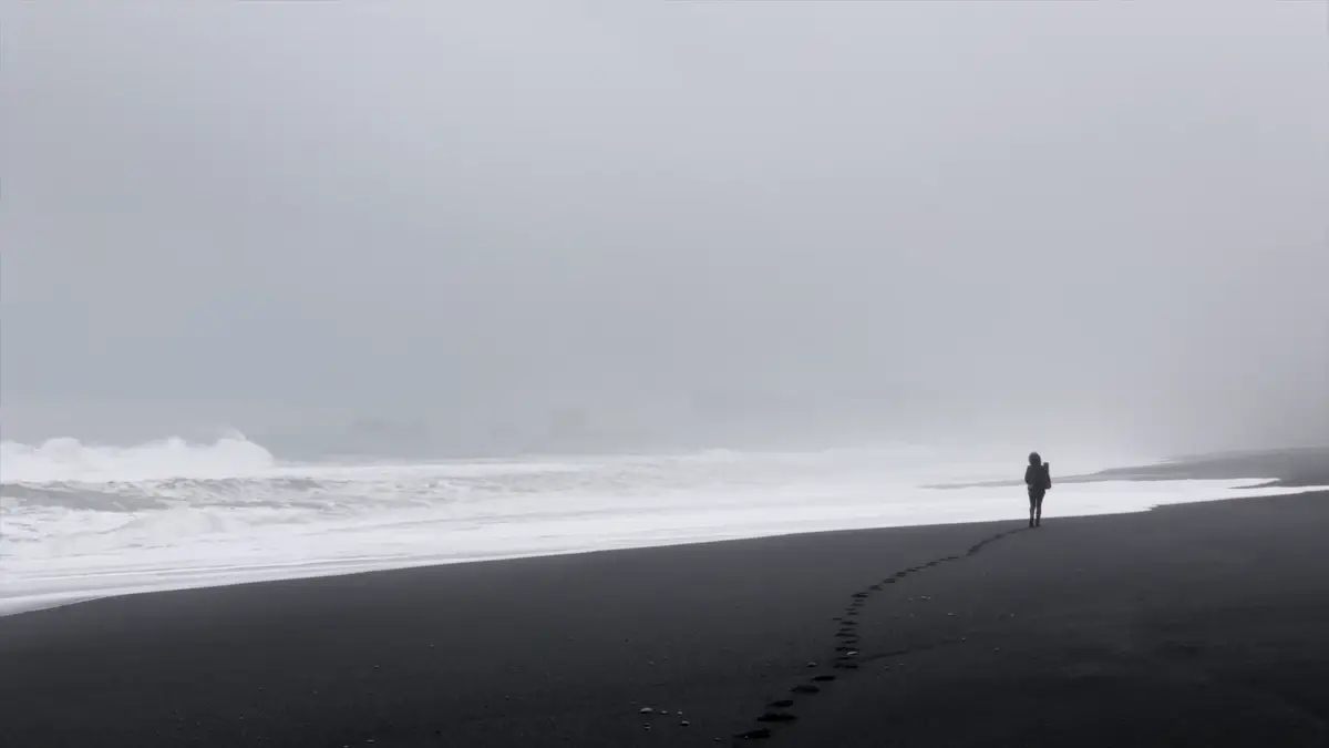 Voyageur marchant seul sur une plage de sable noir en Islande, entouré de brouillard avec l’océan agité en arrière-plan. Un incontournable d’un road trip en camping-car.