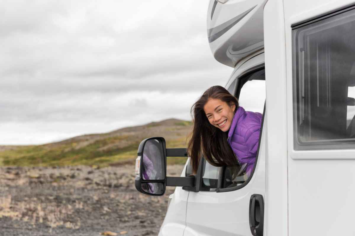 Une femme brune aux cheveux longs sort la t&ecirc;te par la fen&ecirc;tre d'un camping-car en souriant &agrave; la cam&eacute;ra.