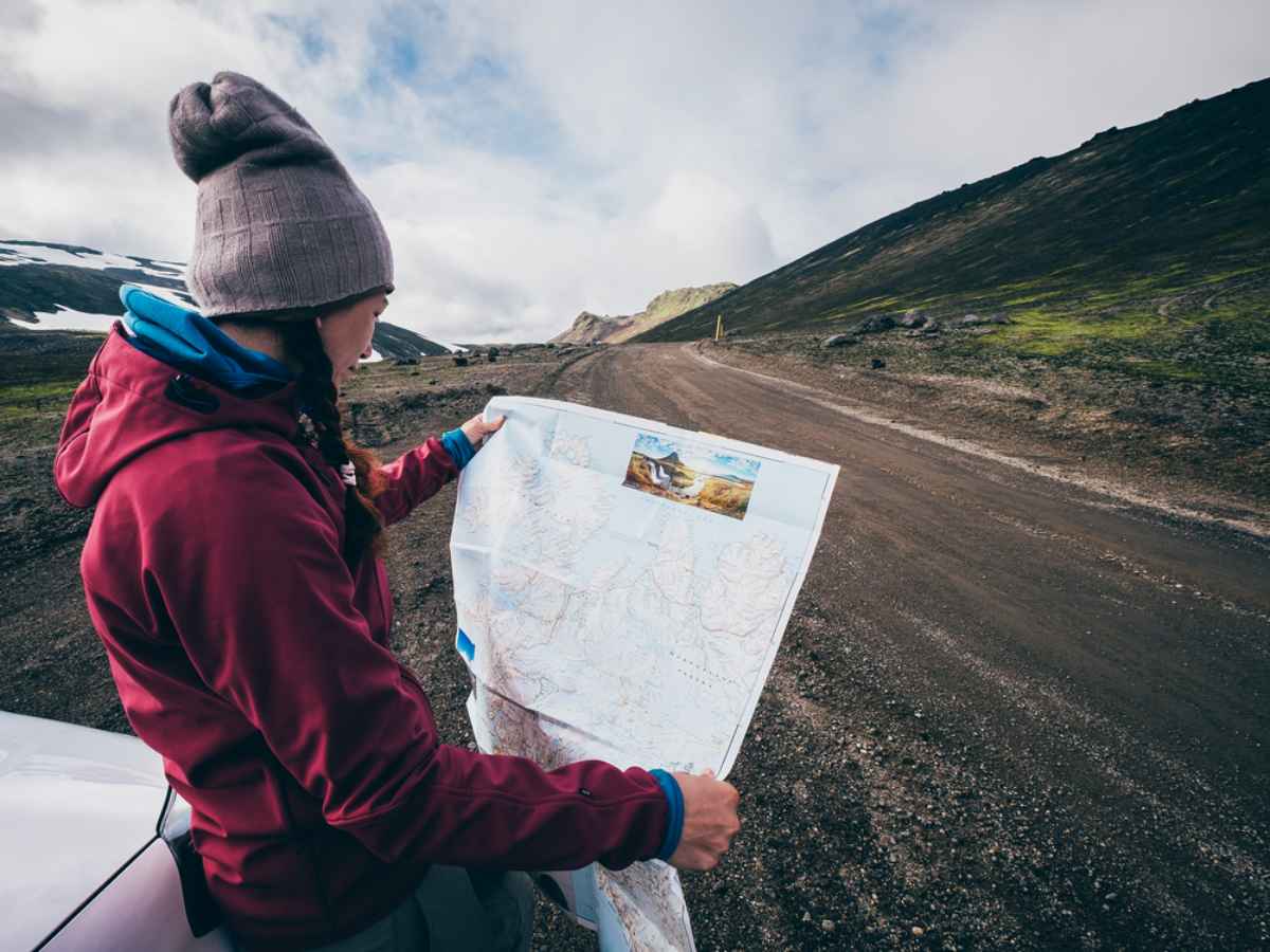 Une femme portant un bonnet et des tresses regarde une carte de l'Islande tandis que son v&eacute;hicule est gar&eacute; sur le bord de la route.