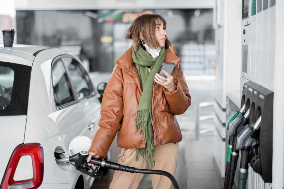 Une fille en manteau de cuir brun tient le pistolet à essence dans sa main droite et l’insère dans le réservoir de son véhicule pour faire le plein, tout en tenant son téléphone de la main gauche pour calculer le prix de l’essence en Islande.