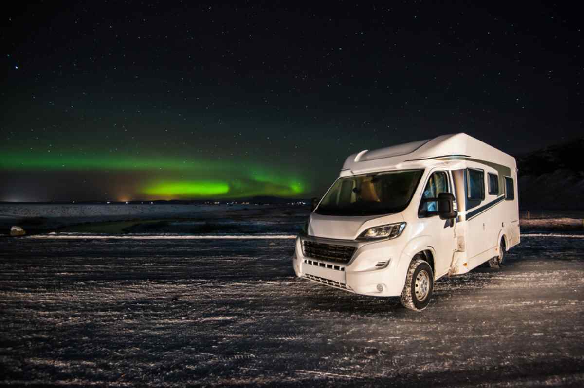 Camping-car gar&eacute; sur la neige sous des aurores bor&eacute;ales en Islande, ciel &eacute;toil&eacute; et lueur verte &agrave; l&rsquo;horizon pour un road trip d&rsquo;hiver.