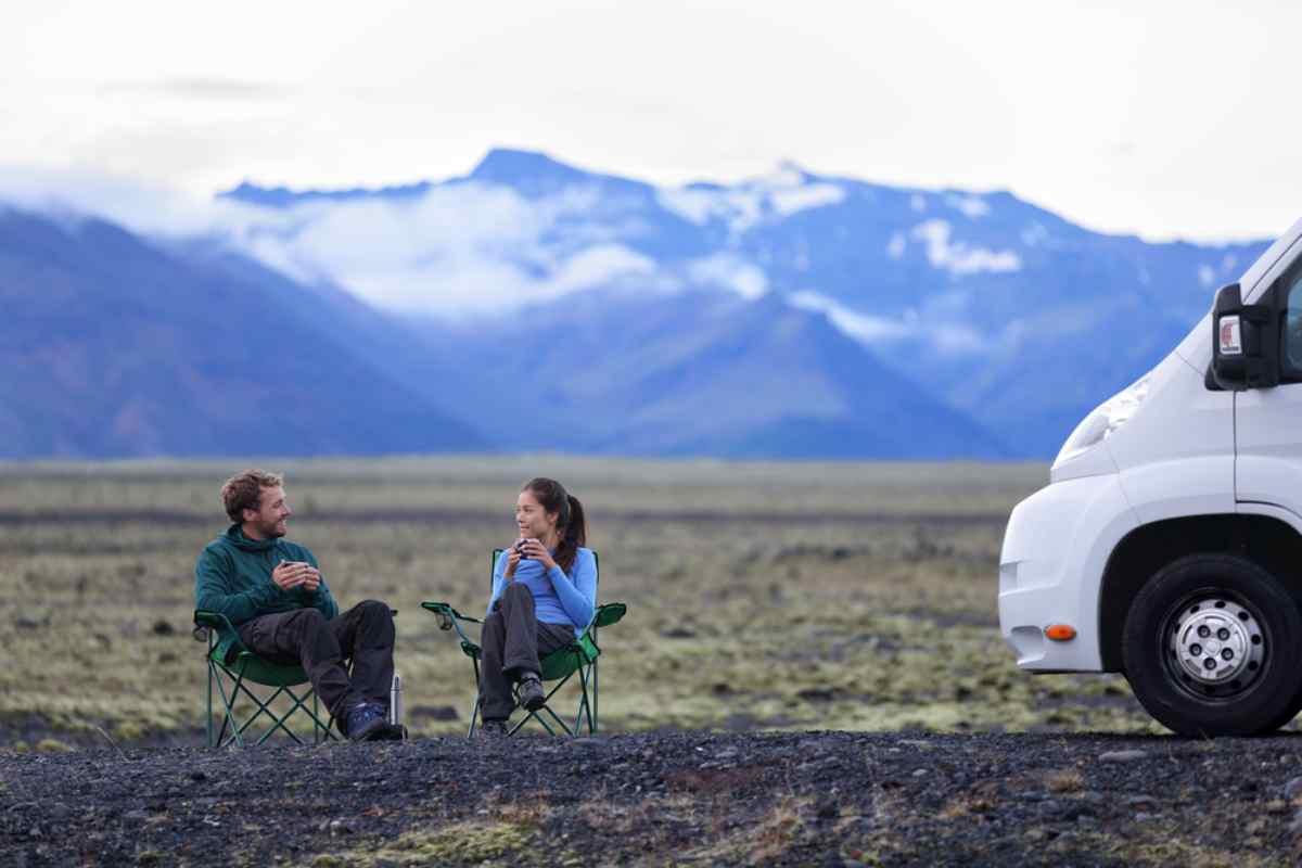 Couple assis sur des chaises pliantes pr&egrave;s d&rsquo;un camping-car; plaine volcanique et montagnes enneig&eacute;es en arri&egrave;re-plan, pause caf&eacute; en route.