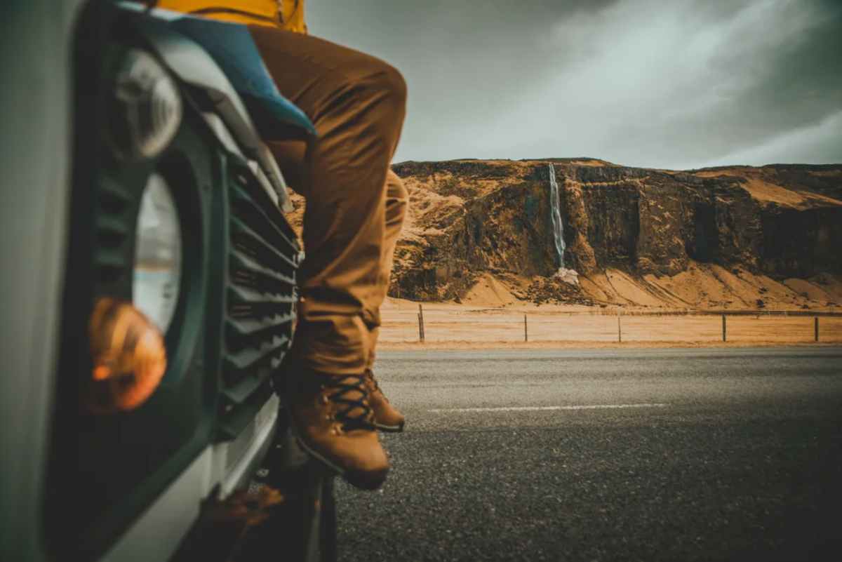 Gros plan sur les jambes d'une personne assise sur le capot de sa voiture garée sur une route islandaise avec une chute d'eau au loin.
